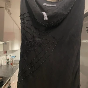 Svart långklänning med pärldetaljer - Elegant svart långklänning med broderade blommor och pärldetaljer i mönster över framsidan. Klänningen har en mjuk draperad halsringning och har tunna band, vilket ger en stilren och sofistikerad look. Perfekt för dig som vill sticka ut med en unik och klassisk stil.