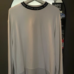 Vit långärmad tröja Acne Studios - Vit långärmad tröja från Acne Studios med svart ribbad krage där varumärket är broderat runt om. Tröjan har en clean och minimalistisk look, perfekt för dig som gillar stilrena plagg med en twist. Mjuk insida och relaxed fit.