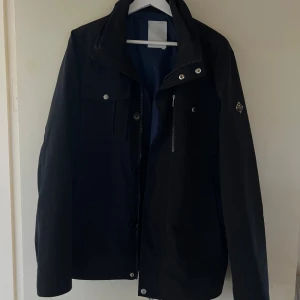 Navy blue field jacket från J.Lindeberg - Stilren navy blå fieldjacket i storlek medium från J.Lindeberg med hög krage, silverfärgade knappar och dragkedja. Jackan har två bröstfickor med lock, emblem på ärmen och justerbara detaljer bak. Perfekt för dig som vill ha en clean look för hösten!