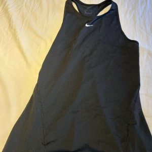 Svart Nike Dri-Fit linnetopp - Säljer en svart sportig linnetopp från Nike med Dri-Fit-teknologi. Toppen har en klassisk racerback-design och ett litet vitt Nike-märke framtill. Perfekt för träning och aktiva dagar, tillverkad i ett lätt och snabbtorkande syntetmaterial.