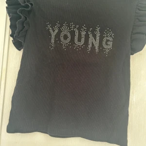 Svart topp med volangärm från Gina Tricot - Svart ribbad topp från Gina Tricot med korta volangärmar och texten 'YOUNG' framtill i silvriga pärlor. T-shirten har en tight passform och är tillverkad i mjukt material som känns skönt mot huden. Perfekt för dig som vill ha en cool och trendig look.