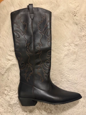Svarta cowboy boots från RAID London - Snygga svarta cowboy boots från RAID London med spetsig tå och låg klack. Boots i skinnimitation med dekorativa bruna sömmar i westernstil längs skaftet. Skorna är endast använda vid ett tillfälle så väldigt fräscha och inga skador:)