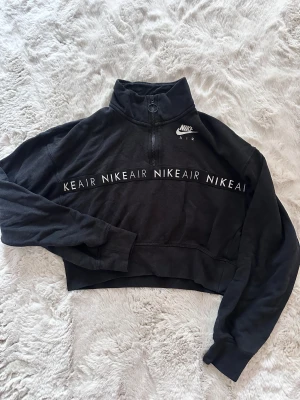 Svart Nike Air croppad half zip tröja - Svart croppad tröja från Nike Air med half zip och hög krage. Snyggt tryck med NIKEAIR över bröstet och logga på vänster sida. Mjuk bomullskänsla och långa ärmar, perfekt för chill eller träning.
