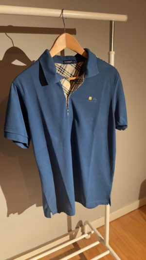 Blå pikétröja Burberry London L - Snygg blå pikétröja från Burberry London i storlek L. Klassisk krage och korta ärmar, med Burberrys ikoniska rutiga mönster på insidan av knappslån. Diskret logga broderad på bröstet. Perfekt för dig som gillar stilrena och tidlösa plagg.