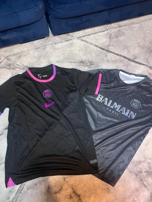 Svarta PSG x Nike & Balmain t-shirts - Två svarta PSG t-shirts, en från Nike med lila och rosa detaljer runt hals och ärmslut, samt PSG-logga och Nike swoosh i lila. Den andra från Balmain med skimrande Balmain Paris-tryck och PSG-logga på bröstet. Båda har rund hals och korta ärmar, tillverkade i polyester.
