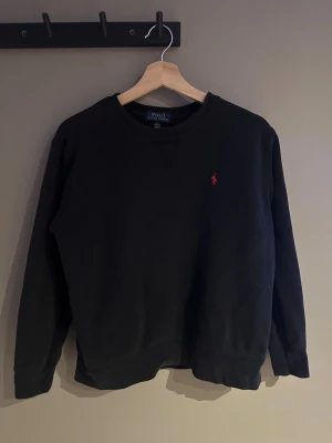 Svart sweatshirt från Polo Ralph Lauren - Svart sweatshirt från Polo Ralph Lauren med klassisk röd broderad logga på bröstet. Tröjan har rund hals, ribbade muddar och är långärmad. Mjuk och skön bomullskvalitet som passar perfekt för chill dagar. Snygg och enkel design som funkar till allt.