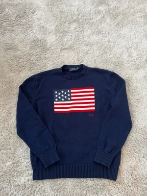 Mörkblå stickad tröja Polo Ralph Lauren - Mörkblå stickad tröja från Polo Ralph Lauren med amerikansk flagga framtill och RL-broderi. Klassisk rund halsringning och ribbade muddar. Perfekt för dig som vill ha en ikonisk och stilren look med en touch av preppy vibes.