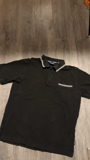 Svart pikétröja från Harvest L - Svart pikétröja från Harvest med klassisk krage och knäppning framtill. Snygga vita ränder på kragen och vid bröstfickan ger en sportig touch. Tillverkad i bomull för skön känsla. Perfekt för dig som gillar stilren och enkel design.