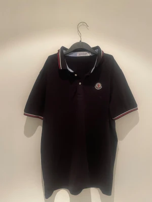 Svart pikétröja från Moncler - Säljer en svart pikétröja från Moncler med klassisk krage och knäppning framtill. Tröjan har Moncler-logga på bröstet och snygga rödvita detaljer på krage och ärmslut. Perfekt för dig som gillar stilrena och sportiga plagg.