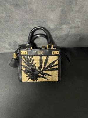 Svart och beige handväska från Guess - Snygg handväska från Guess med svart och beige flätad design, dekorerad med svart broderat blommönster på framsidan. Väskan har svarta skinn-detaljer, guldfärgade metalldelar och dragkedja upptill. Kommer med både handtag och avtagbar axelrem.// den är 20 cm kanske 