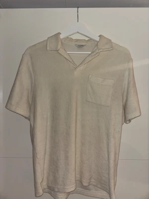 Beige frottéskjorta från East West - Beige kortärmad skjorta i mjuk frotté från East West. Skjortan har klassisk krage, bröstficka och avslappnad passform. Perfekt för chill dagar när du vill ha något bekvämt men ändå stilrent.