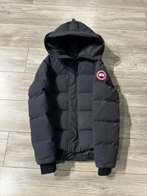 Canada goose Macmillan jacka - Säljer en mörkblå dunjacka från Canada Goose med huva och klassisk logga på ärmen. Jackan har en quiltad design och stängs med dragkedja och knappar. Perfekt för kalla dagar och har en snygg, clean look.