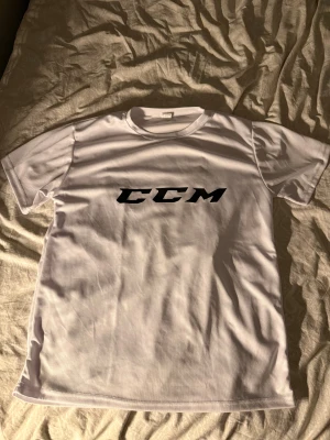 Vit CCM t-shirt i polyester, strl M - Fräsch vit t-shirt från CCM med svart logga tryckt på bröstet. Modellen är klassisk med rund hals och korta ärmar. Materialet är lätt polyester som andas bra, perfekt för sport eller chill. Snygg och enkel design som funkar till det mesta.