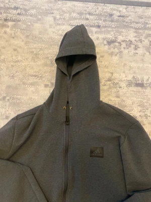 Mörkgrå hoodie från Adidas - Snygg mörkgrå hoodie från Adidas med dragkedja hela vägen och svart logga på bröstet. Modellen har huva med metallöljetter och är tillverkad i mjukt sweatshirtmaterial. Perfekt för en chill och sportig look.
