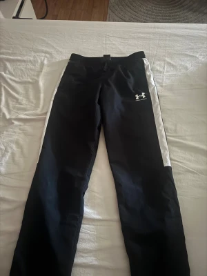 Svarta träningsbyxor från Under Armour - Svarta träningsbyxor från Under Armour med vita sidopaneler och elastisk midja. Byxorna har en sportig look med logga på låret och är tillverkade i ett lätt och ventilerande syntetmaterial. Perfekta för dig som gillar att röra på dig. 