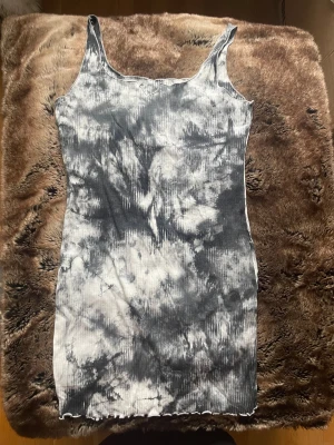 Svartvit ribbad kortklänning - Säljer en svartvit ribbad kortklänning med coolt batikmönster. Klänningen är ärmlös och har en unik detalj vid halsringningen. Materialet är mjukt och stretchigt, perfekt för dig som gillar en avslappnad men trendig stil.