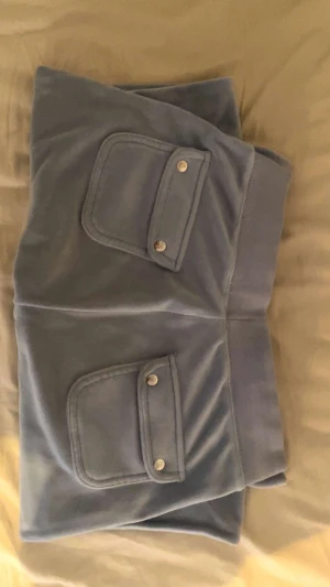 Blå velour Juicy Couture byxor  - Snygga ljusblå shorts från Juicy Couture i mjuk velour. Modellen har låg midja med bred resår och snörning, samt stora fickor framtill med lock och tryckknappar. Perfekta för en chill och trendig look.