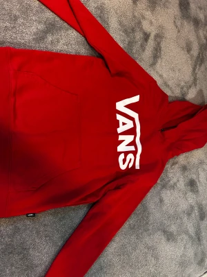Röd hoodie från Vans med logga - Säljer en röd hoodie från Vans i storlek medium. Tröjan har stor vit Vans-logga på bröstet, huva och en klassisk magficka. Materialet är mjuk bomull och passformen är normal. Perfekt för dig som gillar streetwear och vill sticka ut. Väldig bra skick, helt oanvänd