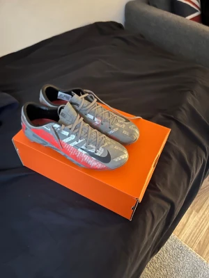 Nike Mercurial grå fotbollsskor - Nike Mercurial fotbollsskor i grått med svarta och röda detaljer. Snyggt mönstrad ovandel med stora reflekterande bokstäver och klassisk svart swoosh. Skorna har snörning och platt sula, insida i rött och svart. Perfekt för dig som vill sticka ut på planen.