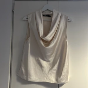 Champagnefärgad satinblus från Zara - Säljer en champagnefärgad ärmlös blus från Zara i glansigt satinmaterial. Toppen har en snygg draperad halsringning och slitsar i sidorna. Perfekt för dig som gillar stilrena och eleganta plagg med en modern twist.
