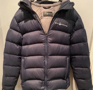 Mörkblå Sail Racing dunjacka - Snygg mörkblå dunjacka från Sail Racing med huva och logga på bröstet. Jackan har en quiltad design, dragkedja framtill och är fodrad i beige. Perfekt för kalla dagar och har en clean, sportig look som funkar till streetwear. Har tre täckta osynliga hål