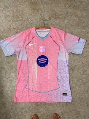 Barça x Nike rosa fotbollströja - Säljer en FC Barcelona x Nike matchtröja i rosa och ljuslila med blå detaljer. Tröjan har korta ärmar, V-ringning och tryck med Spotify-logga samt klubbmärke framtill. Materialet är lätt och andas, perfekt för fotboll eller träning.