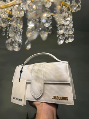 Jacquemus the large bambino - Supersnygg vit handväska från Jacquemus med stilren, rektangulär form och guldfärgad logga. Väskan har ett topphandtag samt avtagbar axelrem med gulddetaljer. Perfekt för dig som gillar lyxig design.