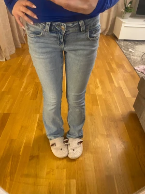 Ljusblå bootcut jeans låg midja - Säljer ett par ljusblå jeans med låg midja och bootcut-snitt. Jeansen har klassisk femficksdesign, slitna detaljer och fransad kant längst ner på benen. Materialet är jeans i bomull och de har dragkedja och knapp framtill.