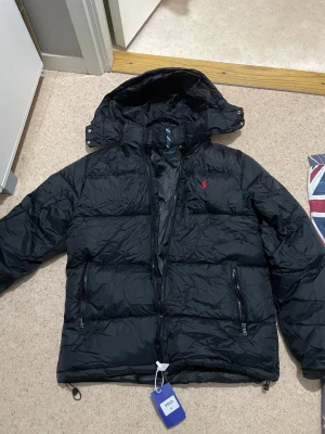 Svart dunjacka från Polo Ralph Lauren - Svart dunjacka från Polo Ralph Lauren med huva och dragkedja. Jackan har två fickor med dragkedja och klassisk röd broderad logga på bröstet. Perfekt för kalla dagar och har en clean, sportig look.