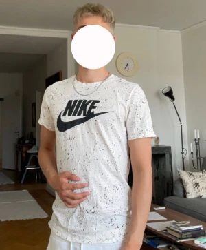 Vit Nike t-shirt med svart logga - Snygg vit t-shirt från Nike med svart logga och coolt stänkmönster över hela tröjan. Modellen är klassisk med rund hals och korta ärmar, perfekt för en sportig och avslappnad stil. Materialet är mjuk bomull som känns skönt mot huden.