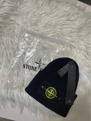 Svart mössa från Stone Island - Ribbstickad svart mössa från Stone Island med klassisk logotyp-patch framtill i gult och grönt. Mössan är i mjukt material och har en uppvikt kant. Perfekt accessoar för att hålla värmen och ge en streetig vibe.
