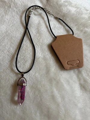 Halsband med rosa kristallhänge - Snyggt halsband med svart snöre och ett spetsigt kristallformat hänge i rosa akryl. Hänget har silverfärgad metallfattning. Perfekt för dig som gillar unika smycken med lite magisk vibe.