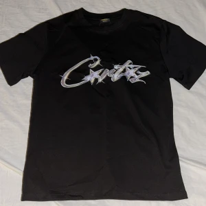 Svart Corteiz t-shirt med metallictryck - Svart t-shirt från Corteiz med ett fett metallictryck över bröstet där det står 'Corteiz' med stjärnor och taggiga detaljer. Klassisk rund hals och korta ärmar. Materialet är mjuk bomull som känns skönt mot huden. Perfekt för dig som gillar streetwear och vill sticka ut.