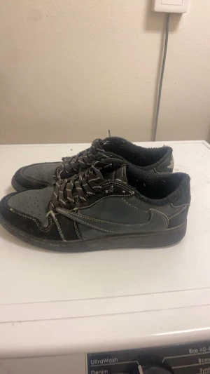 Nike Air Jordan 1 Low svart/grå - Säljer ett par Nike Air Jordan 1 Low i svart och grått med kontrasterande vita sömmar. Skorna har klassisk snörning, perforerad tå och broderade detaljer på hälen. Materialmix av mocka och skinn ger en schysst look. Perfekta för dig som gillar streetwear och sneakers med edge.