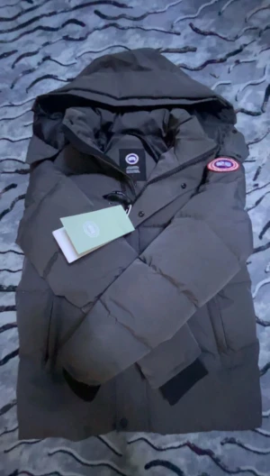 Mörkgrå Canada Goose dunjacka med päls - Säljer en mörkgrå dunjacka från Canada Goose med ikonisk röd logga på ärmen och fluffig beige päls på huvan. Jackan har dragkedja, knappar och ribbade muddar vid ärmslut. Perfekt för kalla vinterdagar och har en snygg, klassisk look.