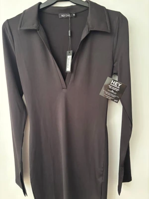 Svart kortklänning med krage från NLY ONE - Svart, stilren kortklänning från NLY ONE med långärmade ärmar och snygg krage. Klänningen har v-ringad hals och slits vid ärmsluten för en cool touch. Materialet är mjukt och stretchigt, vilket gör den bekväm att bära. Perfekt för dig som gillar minimalistisk stil. Oanvända 