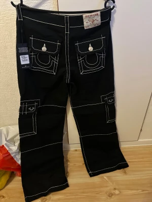 Svarta cargopants från True Religion - Svarta cargopants från True Religion med kontrasterande vita sömmar och stora fickor med lock och silverknappar. Byxorna har bred passform och coola detaljer som extra fickor på benen. Perfekt för dig som gillar streetwear och vill sticka ut. Skriv för fler bilder, och pris kan diskuteras 