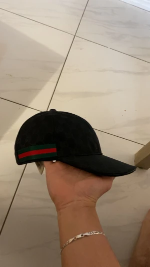 Svart keps från G med GG-mönster - Svart keps från GG med klassiskt GG-monogram i tyget och en grön-röd rand på sidan. Justerbar rem baktill i läder och insida med etikett. Snygg och stilren accessoar som lyfter din outfit.