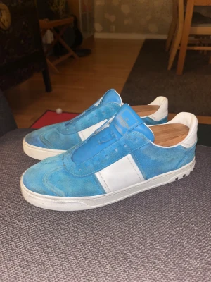Valentino Garavani blå sneakers - Säljer ett par blå och vita Valentino Garavani sneakers i mocka och skinn. Skorna har klassisk låg profil, rund tå, snörning och coola nitar på sulans bakkant. Perfekta för dig som vill sticka ut med en lyxig streetstyle-vibe. 