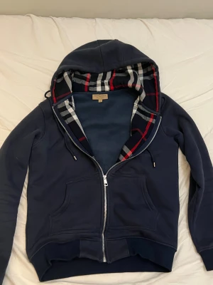 Mörkblå Burberry hoodie med dragkedja - Snygg mörkblå hoodie från Burberry med klassiskt rutigt foder i huvan i svart, vit och röd. Tröjan har dragkedja, snörning i huvan och två stora fickor framtill. Perfekt för dig som gillar stilrena märkesplagg med ikoniska detaljer. Använd endast några gånger väldigt sparsamt.