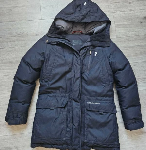 Svart parkajacka Peak Performance S - Svart parkajacka från Peak Performance med huva, flera fickor och dragkedja framtill. Jackan har en stilren look och är perfekt för kalla dagar. Materialet är slitstarkt och jackan har en normal passform. Snygg logga på bröstet och fickan.