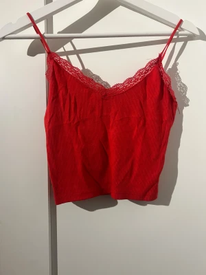 Röd spetskantad linnetopp Stradivarius - Säljer en röd linnetopp från Stradivarius med tunna axelband och spetskant upptill. Toppen är ribbad och har en croppad passform som ger en trendig look. Perfekt för att lyfta vilken outfit som helst med sin klara färg och fina spetsdetaljer.