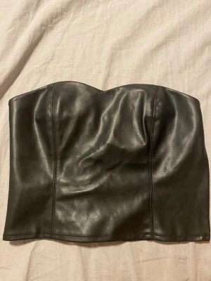 Svart skinn tubtopp med smock - Säljer en svart tubtopp i skinnmaterial. Baksidan har smockdetalj för extra stretch och bekväm passform. Den är i storlek xs