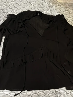 Svart elegant volangblus  - Svart blus från SHEIN i storlek XS med långa ärmar. Blusen har volangdetaljer längs axlar och framtill samt knytband vid halsen. Tunt, lätt material som ger en luftig känsla och snygg passform.