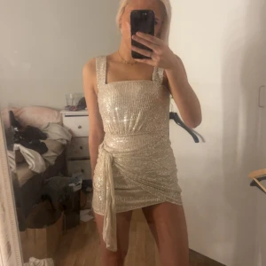 Champagne/guldfärgad klänning - Kortklänning med glittriga paljetterKlänningen har en snygg omlott-detalj med knyt till sidan. Perfekt till nyår eller något annat event. Går att dra ner och upp kjolen till en viss längd. Ingen aning vart den är ifrån tror den är köpt utomlands. 💛