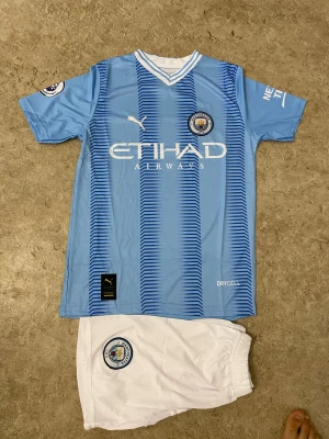 Manchester City Haaland #9 matchtröja - Säljer en ljusblå Manchester City t-shirt från Puma med Haaland och nummer 9 på ryggen. Tröjan har korta ärmar, vitt och blått randigt mönster framtill, klubbmärke och sponsortryck. Materialet är polyester med Drycell-teknologi för att hålla dig torr under fotbollsmatcher.