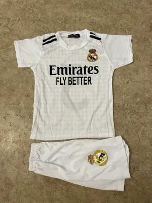 Real Madrid Bellingham #5 t-shirt - Säljer en vit Real Madrid t-shirt med Bellingham och nummer 5 på ryggen. Tröjan har korta ärmar, svarta ränder på axlarna och klubbens emblem på bröstet. Materialet är lätt polyester, perfekt för fotboll. Emirates Fly Better tryck på framsidan.