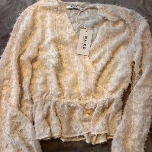 Vit omlottblus med struktur från NA-KD - Säljer en vit omlottblus från NA-KD i storlek 38. Blusen har lång ärm, v-ringning och ett transparent tyg med snyggt strukturerat mönster. Midjan är lätt rynkad och nederdelen har en liten volang. Perfekt för dig som gillar trendiga detaljer och ljusa toner.