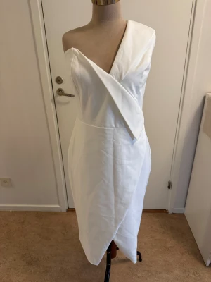 Vit oneshoulder klänning Lavish Alice - Snygg vit klänning från Lavish Alice med asymmetrisk oneshoulder-design och omlott framtill. Klänningen har en figurnära passform och stängs med dragkedja bak. Perfekt för dig som vill sticka ut med en stilren och modern look.