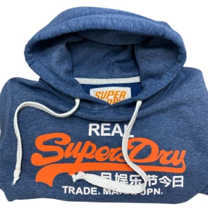 Blå Superdry hoodie  - Blå hoodie från Superdry med stort orange och vitt tryck på bröstet. Tröjans storlek är M, men passar XS Bättre.